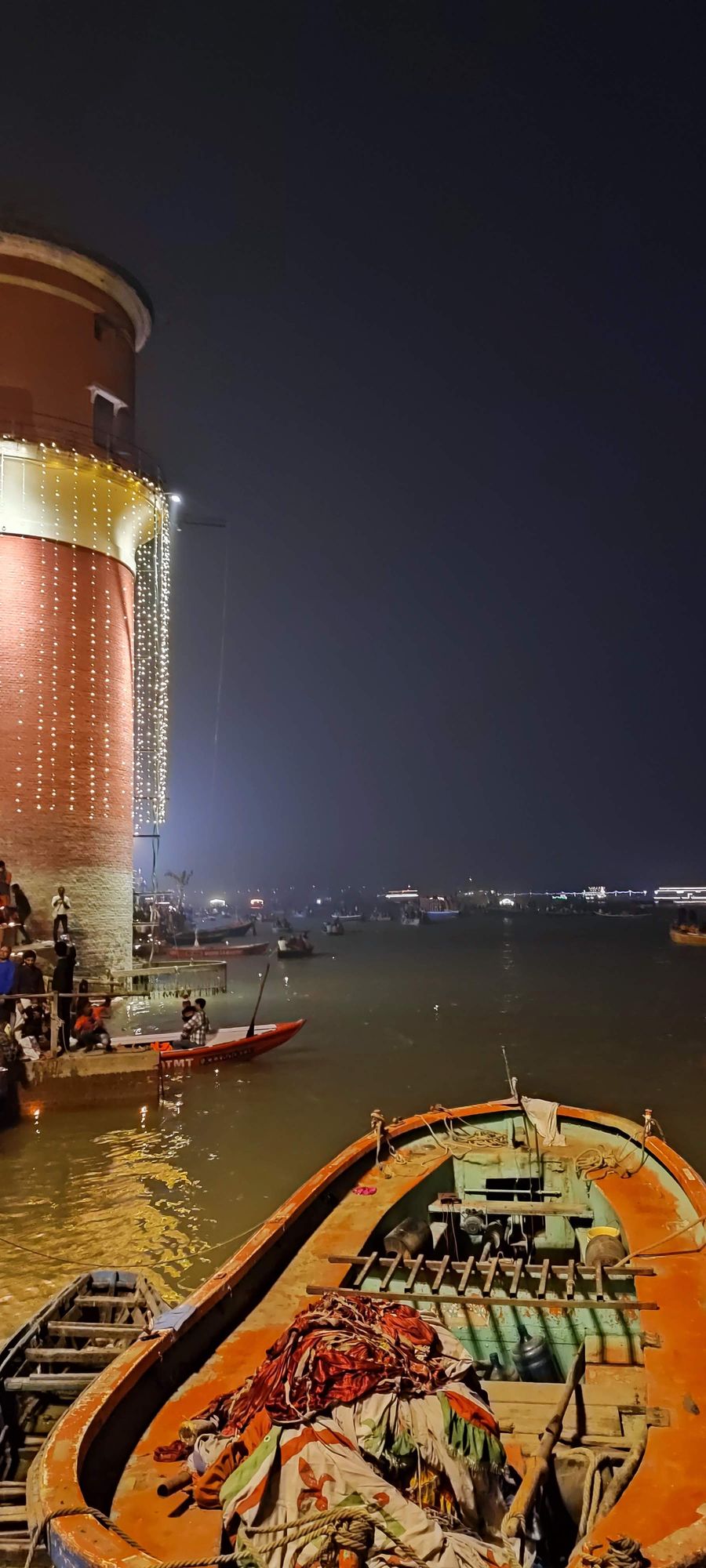 Night at a ghat in Banaras: a brick tower strung with lights, boats on the Ganga